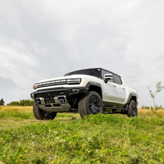 White Hummer EV parked in a natural landscape, showcasing its rugged design and off-road capabilities with grass and wildflowers in the foreground under a cloudy sky.
