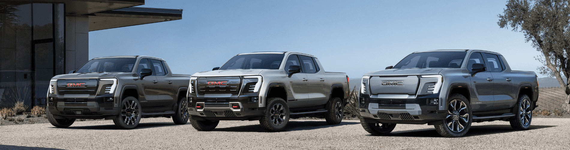 Three new GMC trucks showcased in a modern outdoor setting, emphasizing their sleek design and powerful stance. The image highlights the vehicles' distinct front grilles and bold lines, set against a clear blue sky and minimalist architecture.