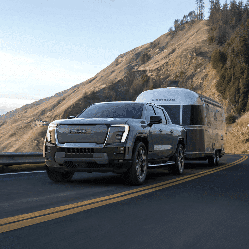 GMC electric truck towing an Airstream trailer on a scenic mountain road, showcasing outdoor adventure and modern vehicle capabilities.