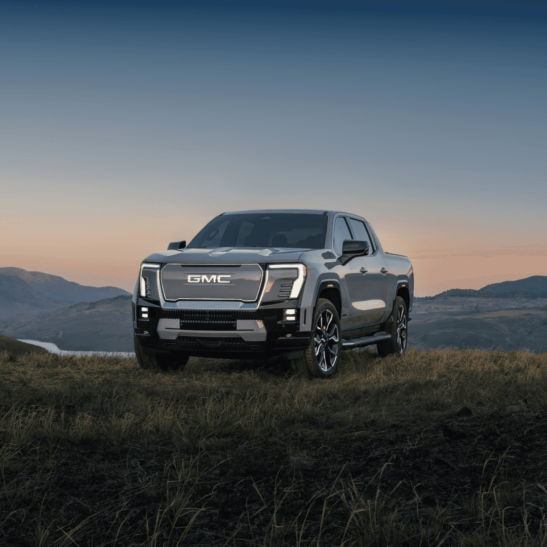 A GMC pickup truck parked on a hillside during sunset, showcasing its modern design, bold front grille, and advanced lighting features against a scenic backdrop of mountains and valleys.