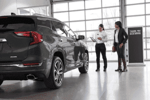 A customer and a sales representative discuss features of a black SUV in a modern car dealership, with large windows and a "Thank You for Your Service" sign in the background.