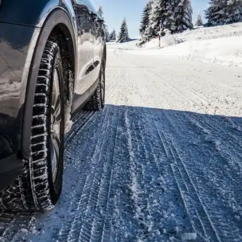 Close-up of a winter tire on a vehicle, showing tread pattern on a snow-covered road surrounded by evergreen trees under clear blue skies, highlighting winter driving conditions.