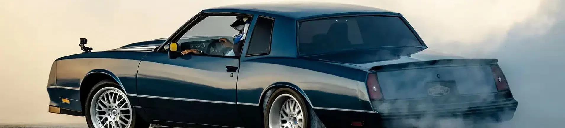 Black muscle car performing a burnout on a racing track, generating smoke and showcasing its powerful rear-wheel drive capabilities. The vehicle features stylish alloy wheels and an aerodynamic body, emphasizing its performance-focused design.