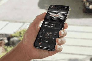 A person holding a smartphone displaying a mobile app interface, featuring the GMC logo and various control options for vehicle functions, set against an outdoor background.