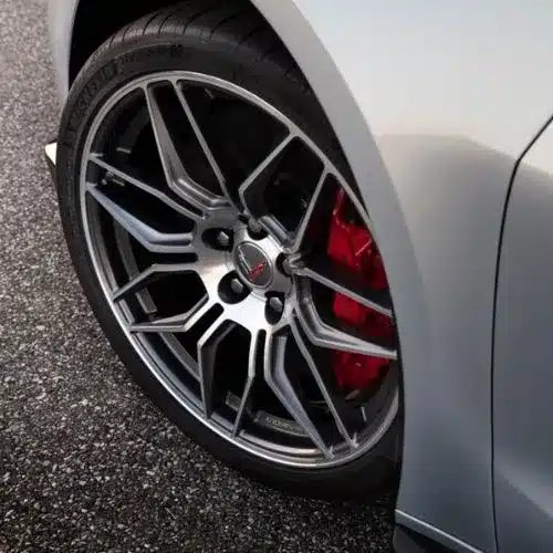 Close-up view of a sleek silver car wheel featuring a multi-spoke alloy design and red brake calipers, set against a textured asphalt background.