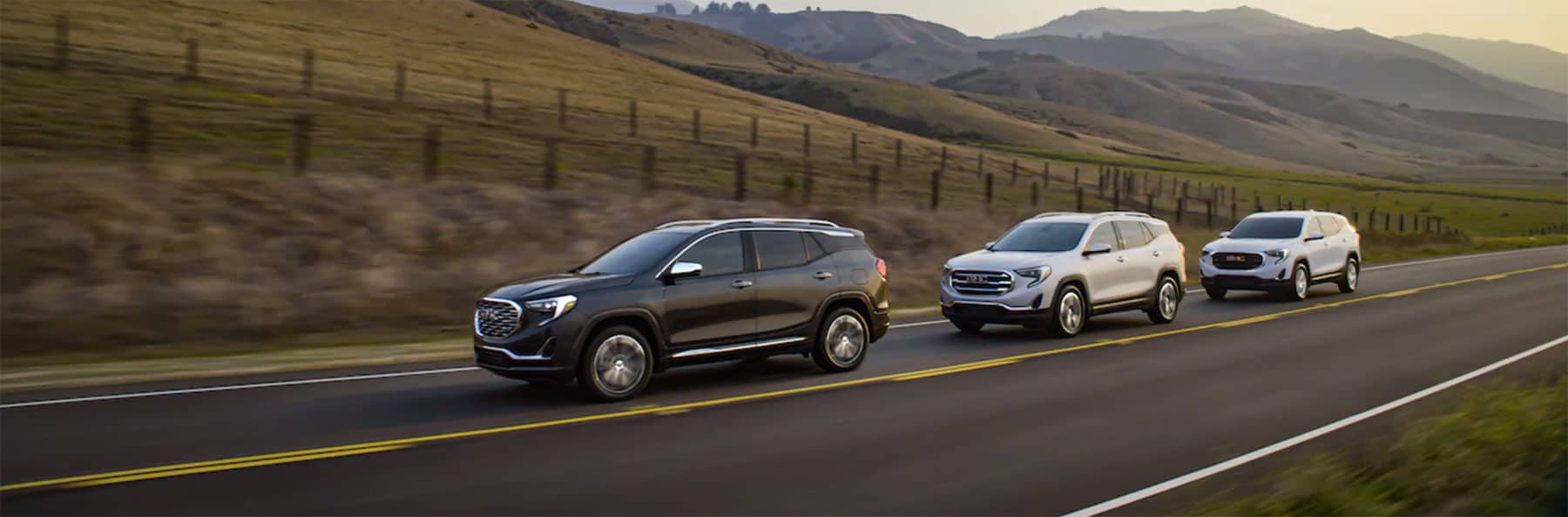 Three GMC SUVs driving along a scenic highway with rolling hills in the background, showcasing modern design and versatility for family adventures.
