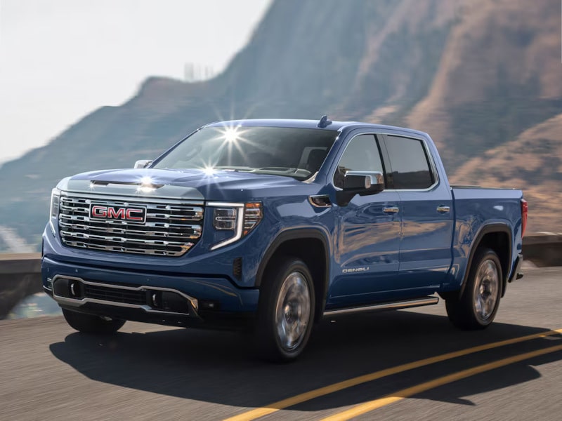 Blue GMC Sierra Denali driving on a winding road with a mountainous background, showcasing its modern design and robust features.