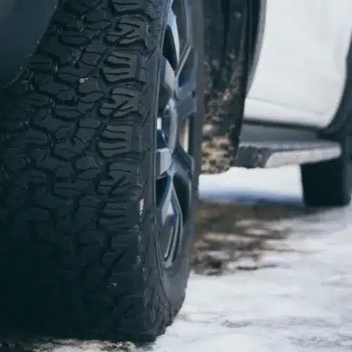 Close-up of a rugged black tire with a detailed tread pattern on a snowy surface, highlighting its winter driving capabilities.