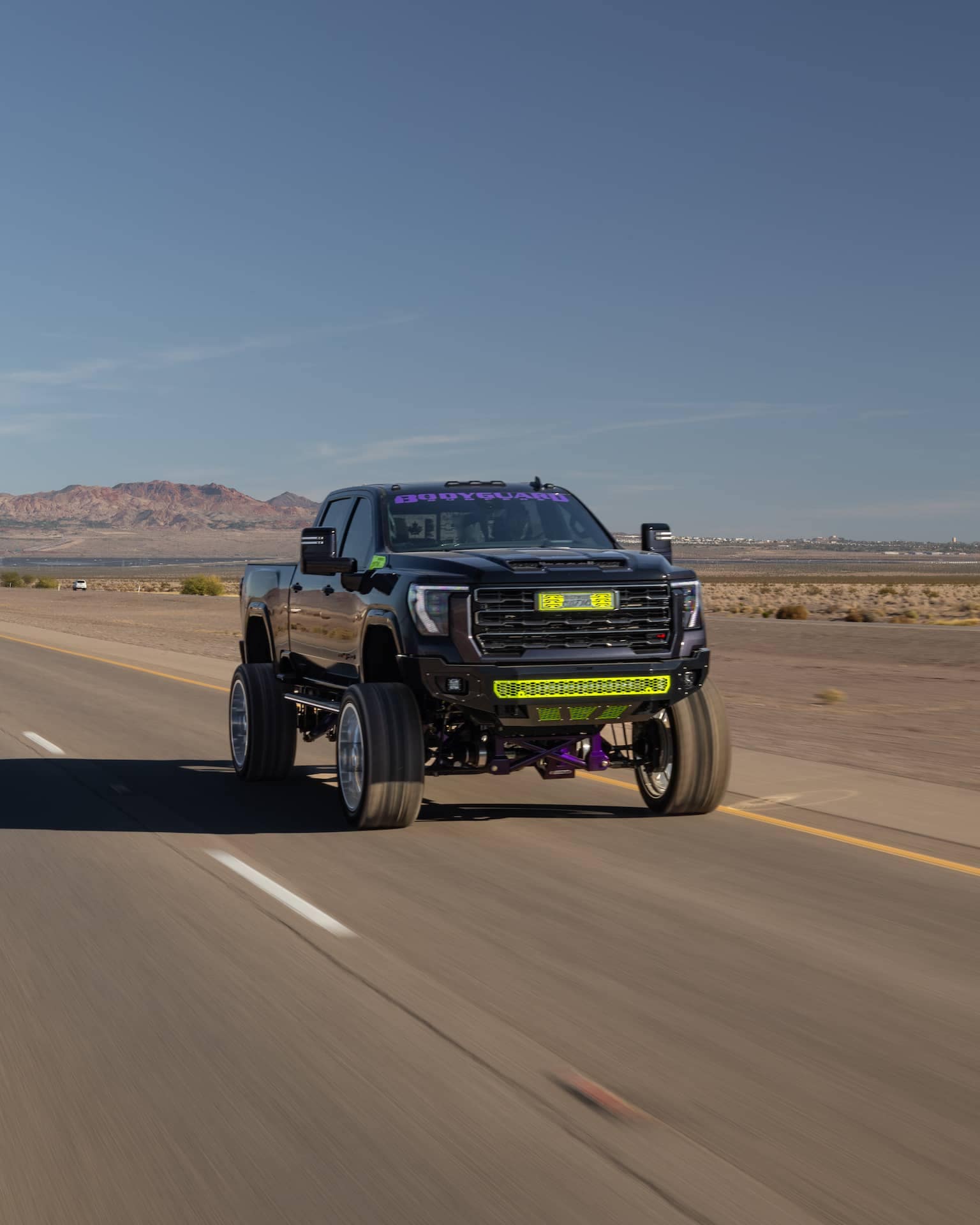 Black lifted truck driving on an open highway with mountain backdrop, featuring vibrant green lights and custom modifications for off-road performance.