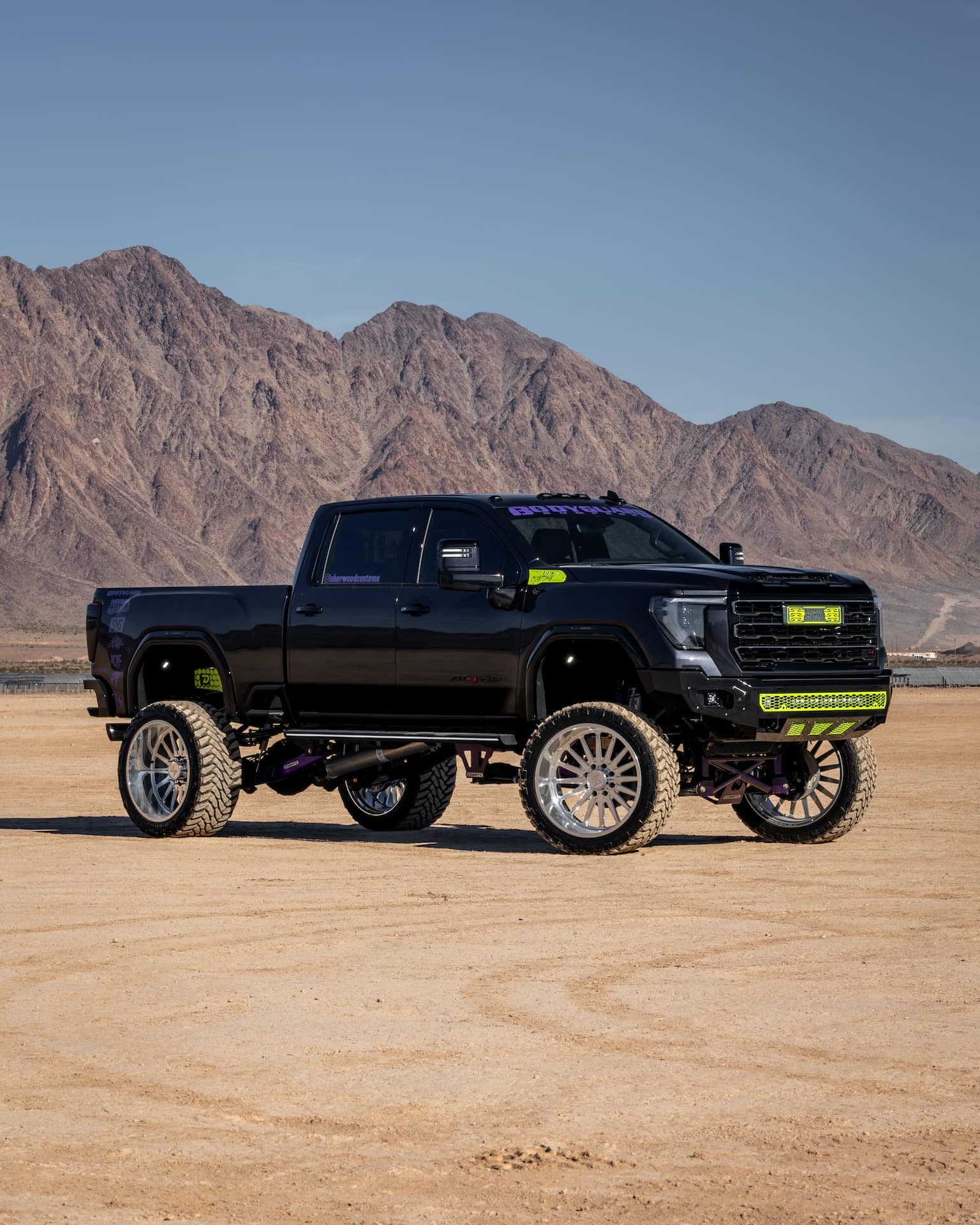 Black lifted truck with oversized wheels parked on sandy terrain, surrounded by rocky mountains under a clear blue sky.