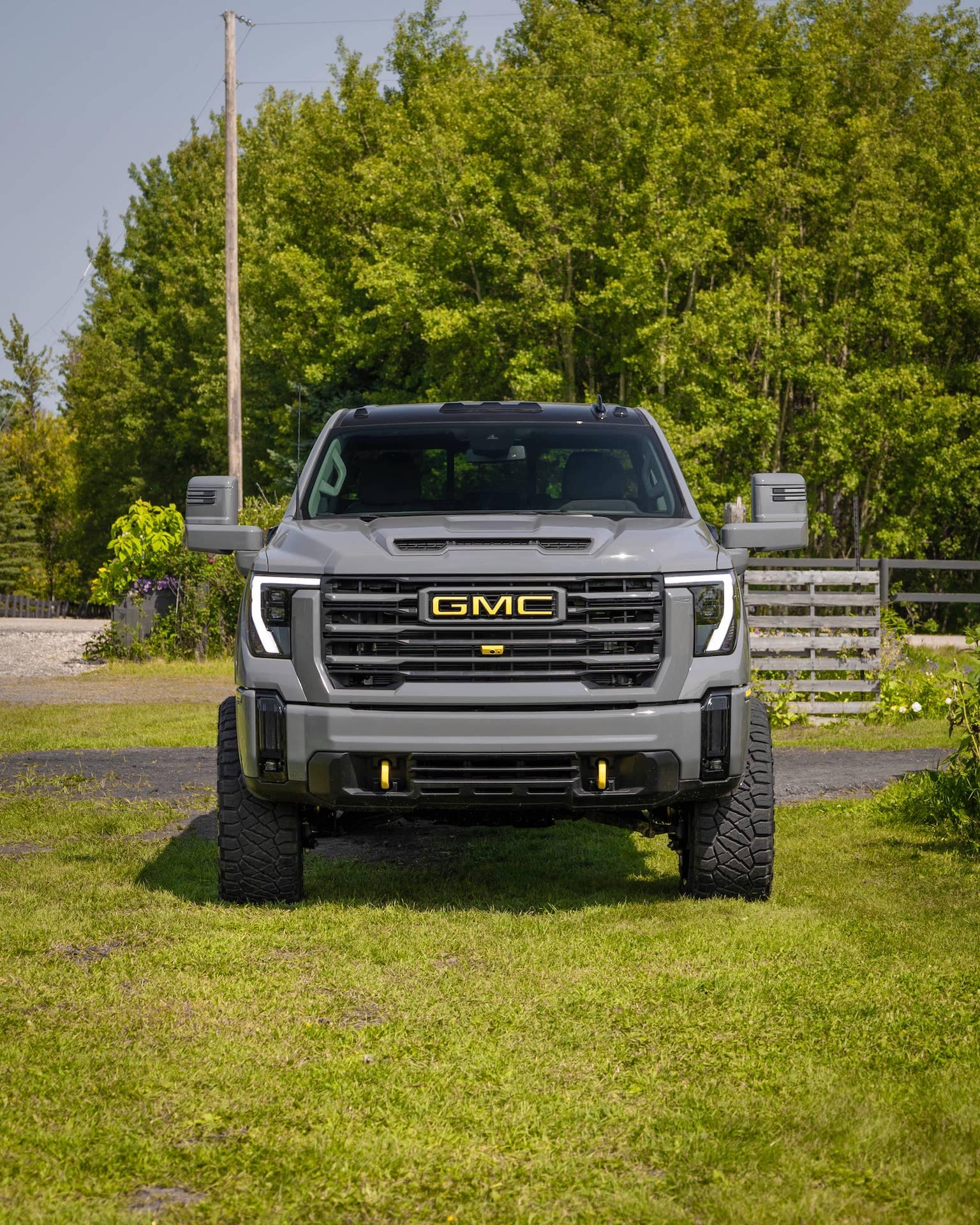 Front view of a lifted gray GMC truck parked on green grass, showcasing its bold design, prominent GMC logo, and rugged tires, surrounded by trees and natural scenery.