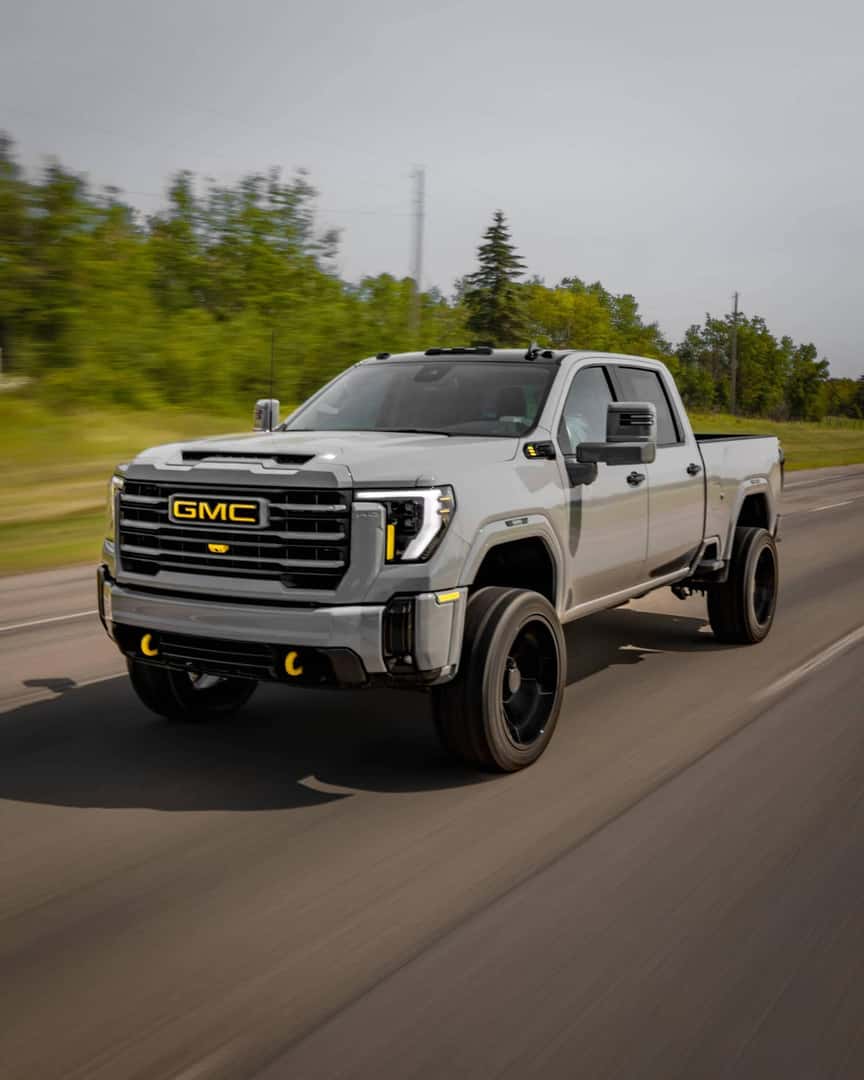 GMC truck driving on a highway with a sleek silver exterior and custom black wheels, showcasing its rugged design and powerful presence on the road.
