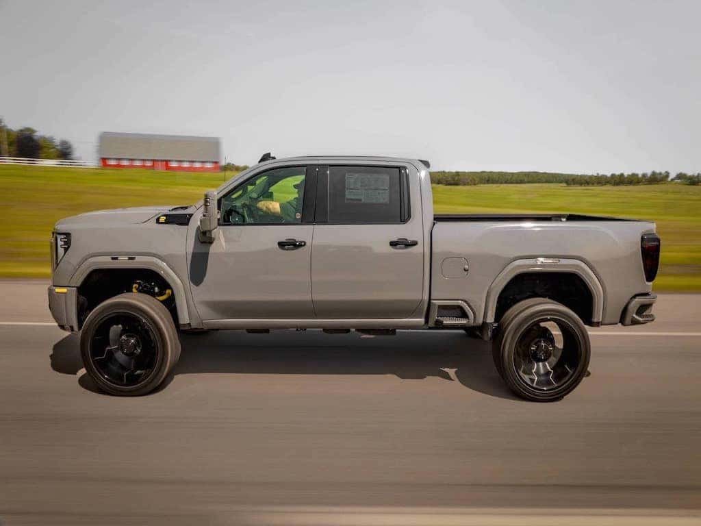 Gray lifted pickup truck driving on a highway with a blurred green landscape in the background, showcasing its oversized black wheels and modern design.