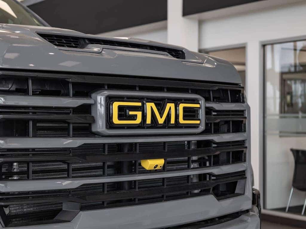 Close-up of a GMC vehicle grille featuring the iconic yellow GMC logo, set against a modern dealership background, showcasing the truck's rugged design and branding.