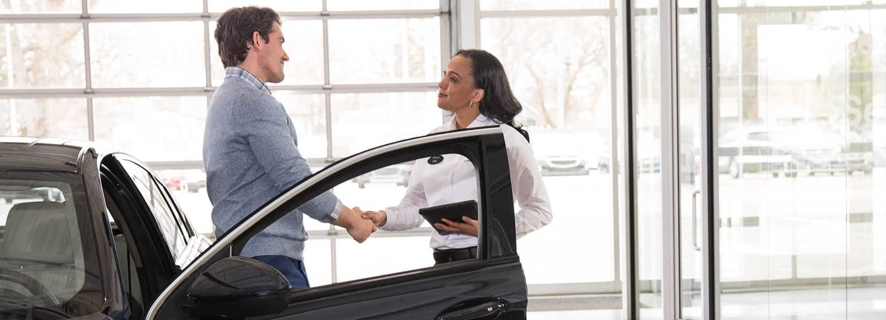 A car salesman and a female customer shake hands inside a modern car dealership, with a vehicle door open nearby, illustrating a successful negotiation and customer service experience.
