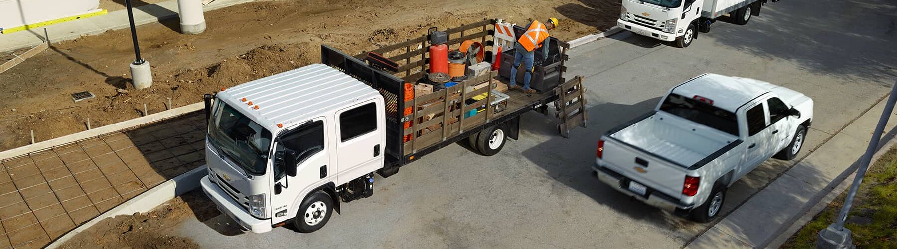 Construction scene featuring a white flatbed truck loaded with tools and materials, parked on a gravel site. Workers in safety gear are engaged in construction activities, while additional white trucks are visible in the background. This image emphasizes construction logistics and teamwork.