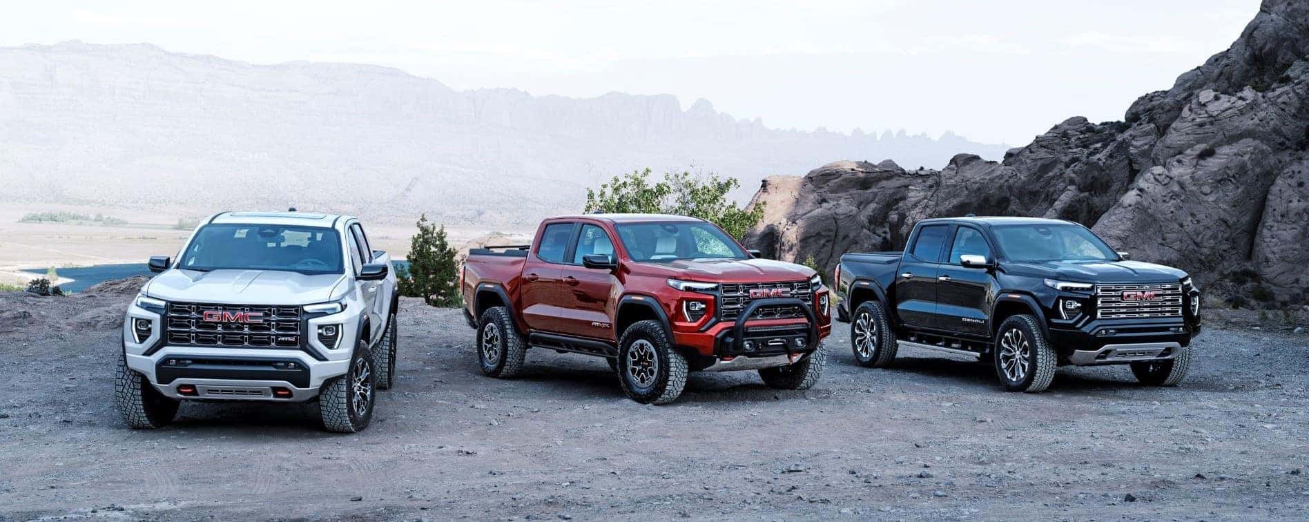 Three GMC trucks parked on rugged terrain, showcasing a white model on the left, a red model in the center, and a black model on the right, against a backdrop of mountains and clear sky. Ideal for off-road adventures and heavy-duty tasks.