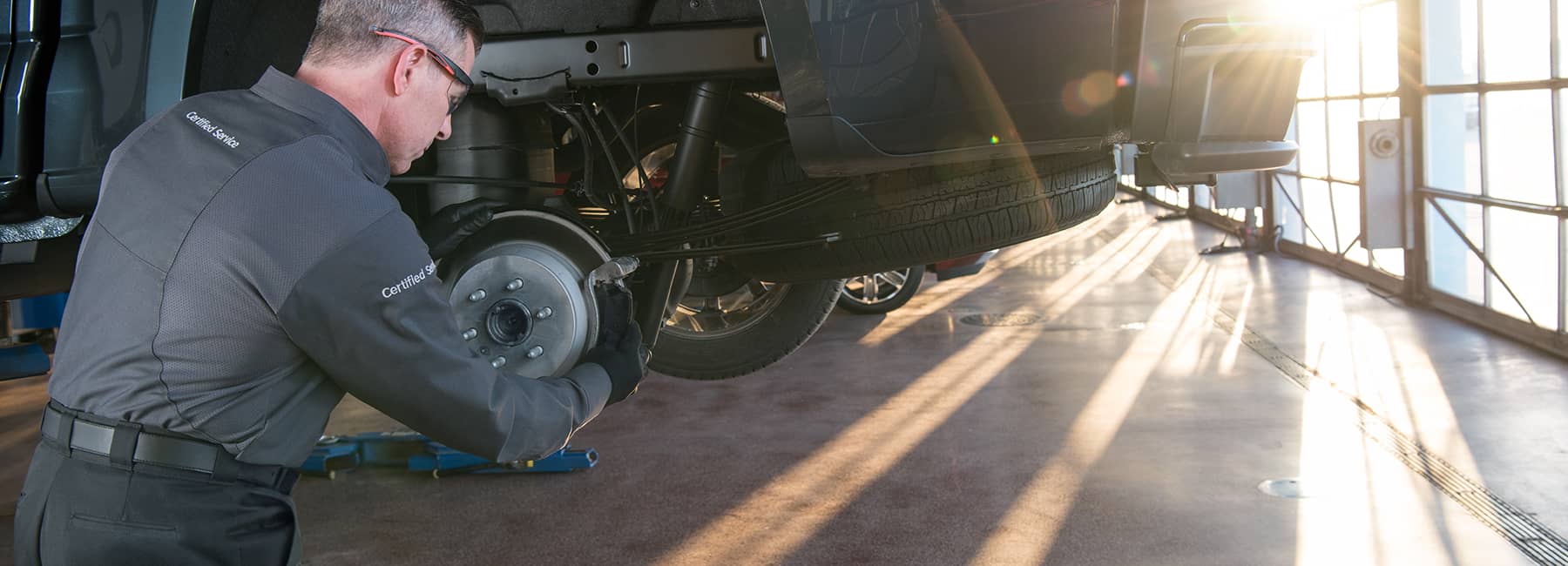 A certified mechanic working on a vehicle's brake assembly in a well-lit garage. Sunlight streams through windows, highlighting the detailed work on the brake drum.