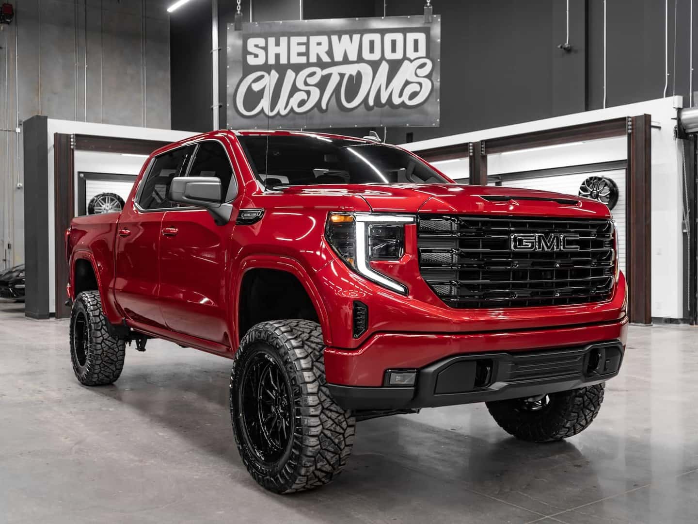 Red GMC truck with a lifted suspension and off-road tires, showcased in a custom vehicle garage with a Sherwood Customs sign in the background.