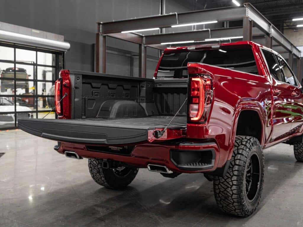 Red pickup truck with an open bed, showcasing a durable black bed liner, set in an industrial garage environment. The truck features off-road tires and modern taillights, highlighting its rugged design and functionality.