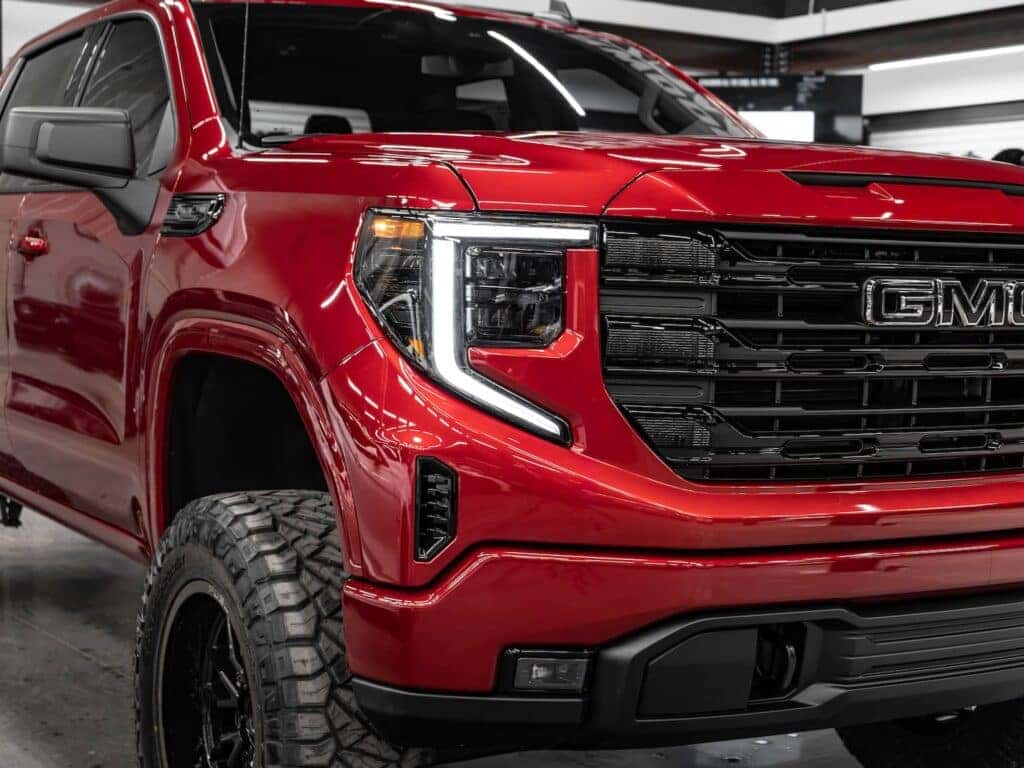 Close-up of a red GMC truck showcasing its aggressive front grille, LED headlights, and off-road tires, highlighting its rugged design and modern features in a showroom setting.