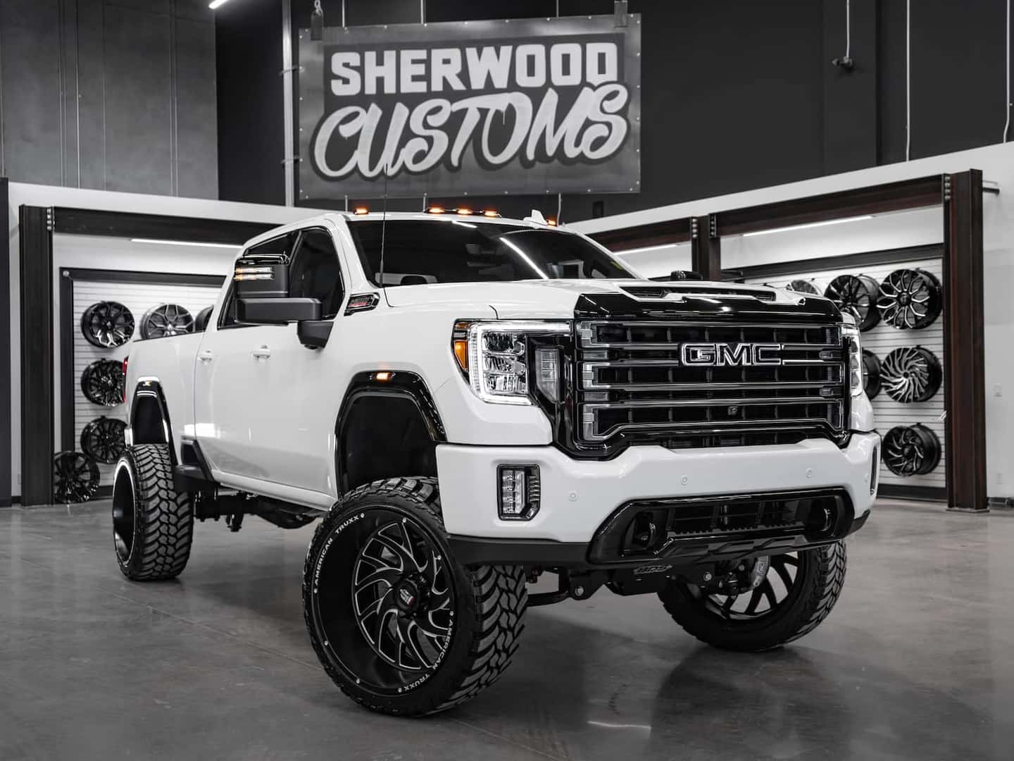 Custom lifted GMC truck in a showroom setting, featuring oversized black alloy wheels and aggressive tires. The vehicle is prominently displayed against a backdrop of custom rims, showcasing its enhanced design and detailing. Ideal for truck enthusiasts and custom vehicle fans.
