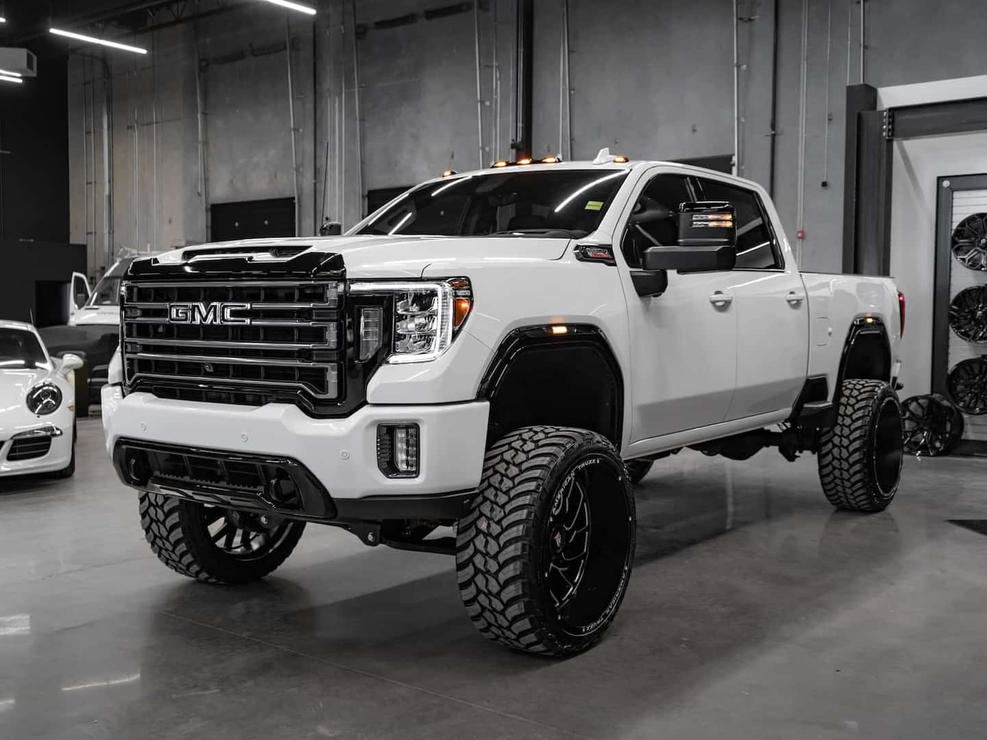 White GMC truck with oversized tires and lifted suspension displayed in a modern showroom, showcasing its rugged design and prominent front grille.