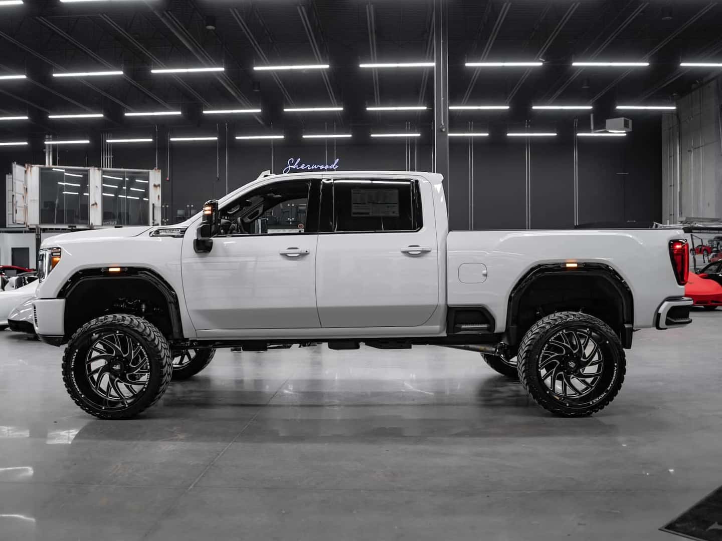 White lifted pickup truck with custom black wheels and an aggressive stance, parked indoors under bright lights. The vehicle features a spacious cab and modern design elements, showcasing its off-road capability and stylish appeal.
