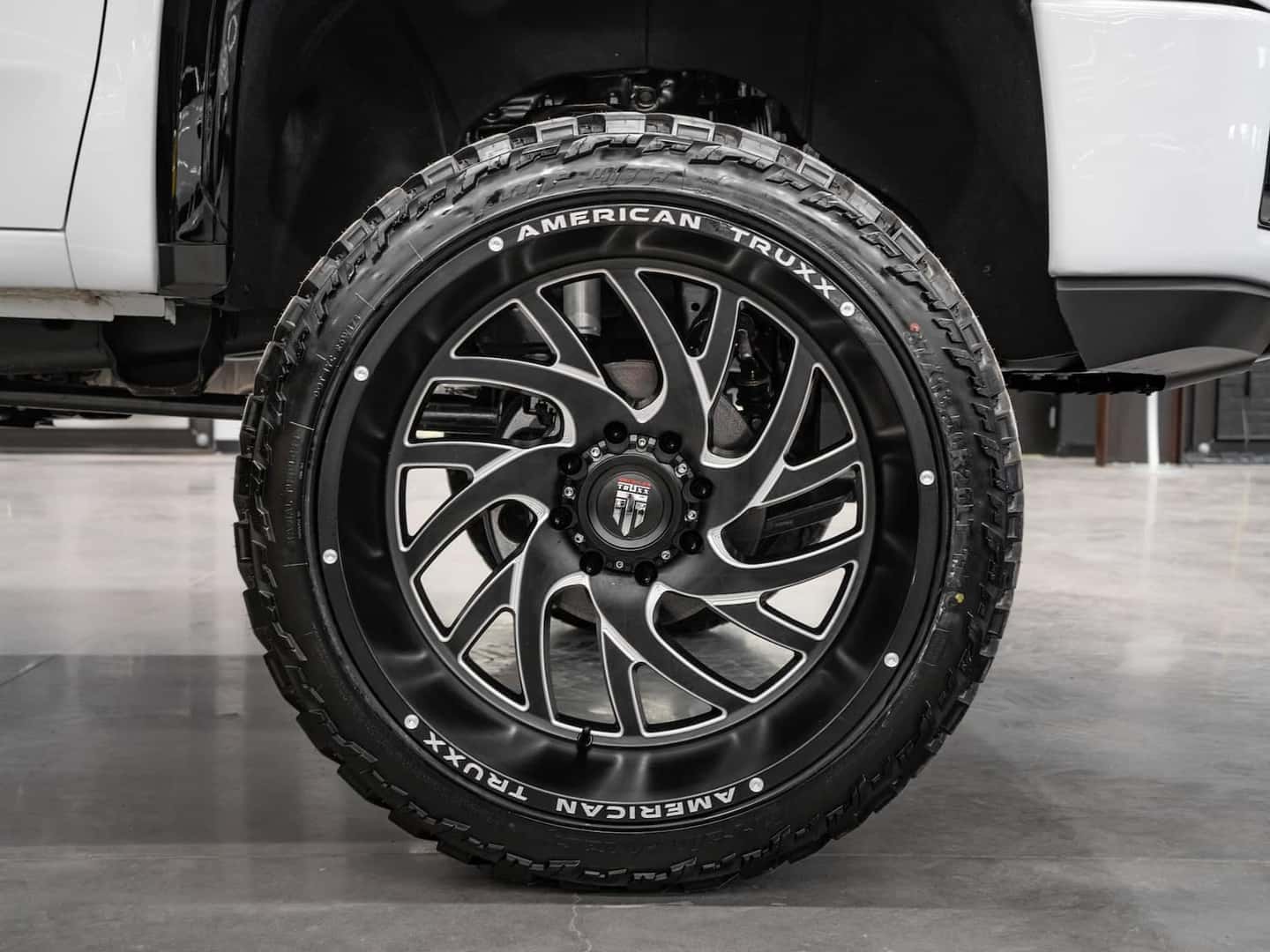 Black alloy wheel with modern design mounted on a truck, featuring a rugged tire from American Truxx brand. The wheel showcases a polished finish and intricate spoke pattern, emphasizing performance and style in off-road vehicles.