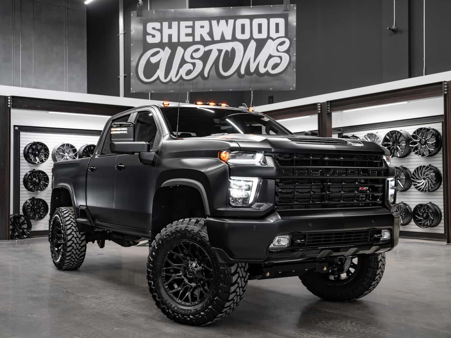 Custom lifted Chevrolet truck showcased in a modern customization shop, featuring a matte black finish, aggressive off-road tires, and stylish black wheels, surrounded by various wheel designs on display.