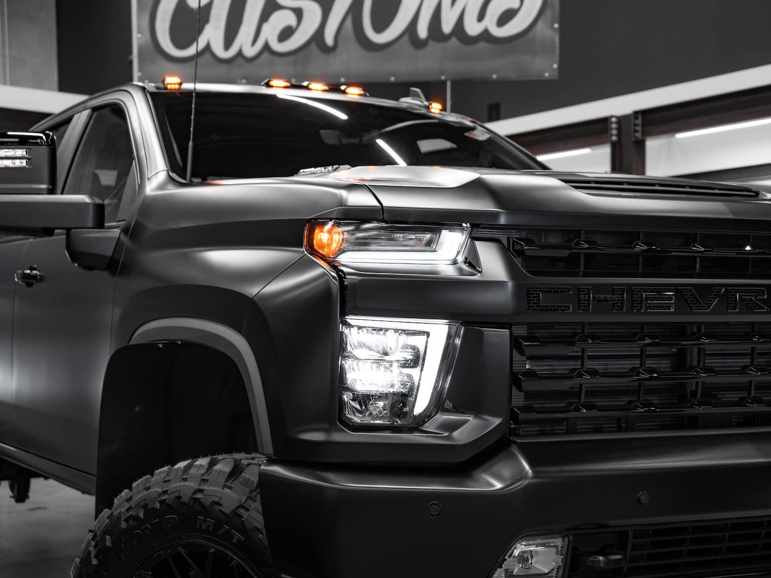 Close-up of a custom matte black Chevrolet truck showcasing its bold front grille, sleek LED headlights, and aggressive design. The image highlights the truck's rugged tires and contrasting accents, set against a modern automotive backdrop. Perfect for showcasing vehicle customizations and aesthetics.