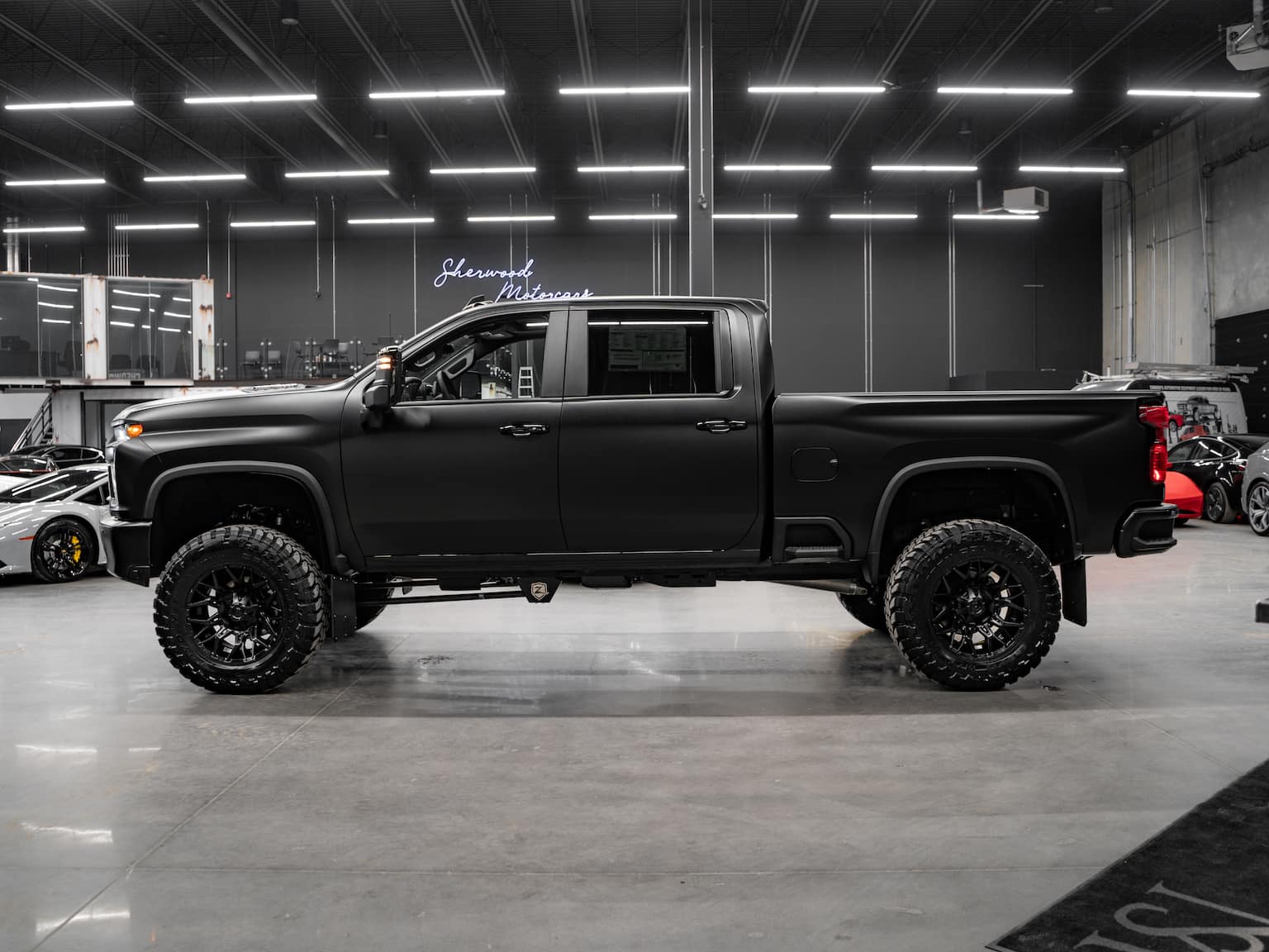 Matte black lifted pickup truck displayed in an automotive showroom, featuring large off-road tires and custom wheels, with luxury sports cars in the background. Ideal for showcasing rugged style and performance enhancements.