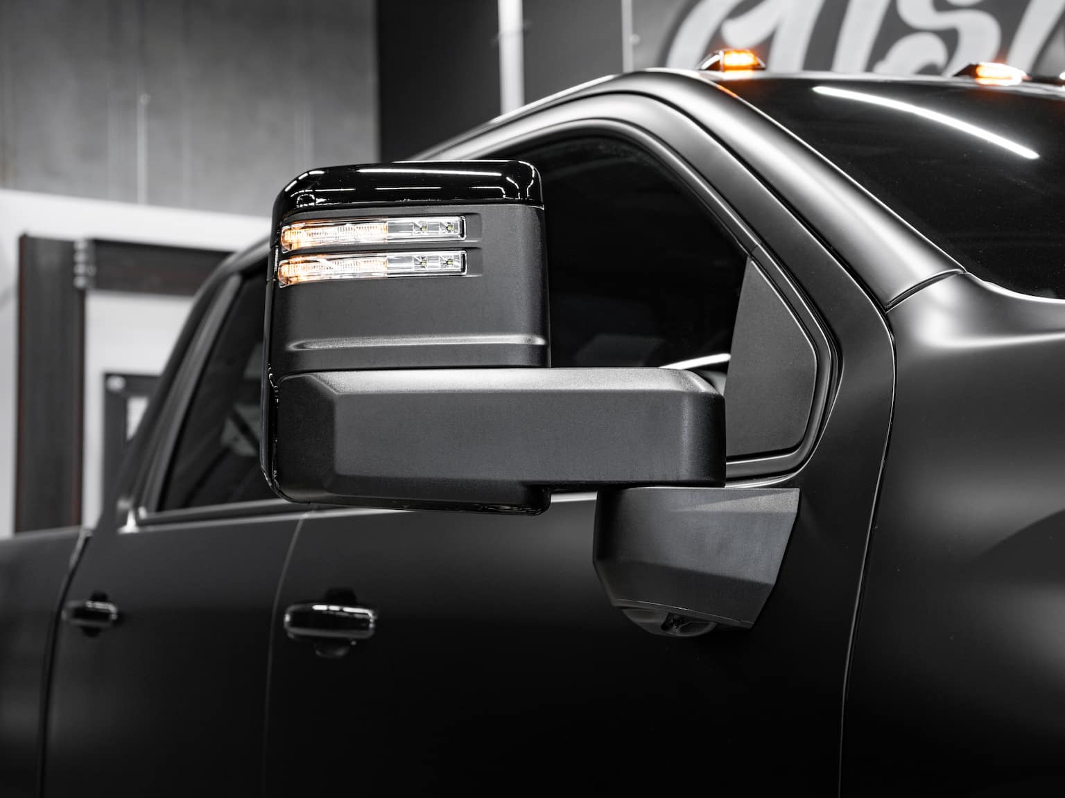 Close-up of a modern truck side mirror with integrated LED lights, showcasing a sleek black finish and detailing, set against a blurred industrial backdrop.