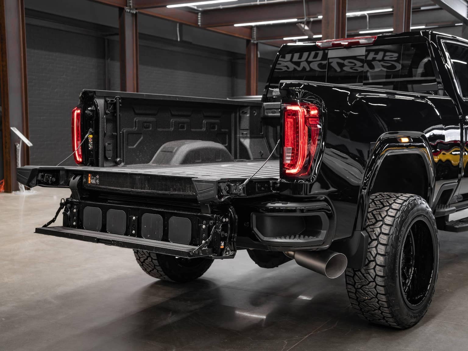 Close-up view of the rear of a black pickup truck, showcasing the open tailgate, bed liner, and custom wheels, emphasizing its rugged design and functionality within an industrial setting.