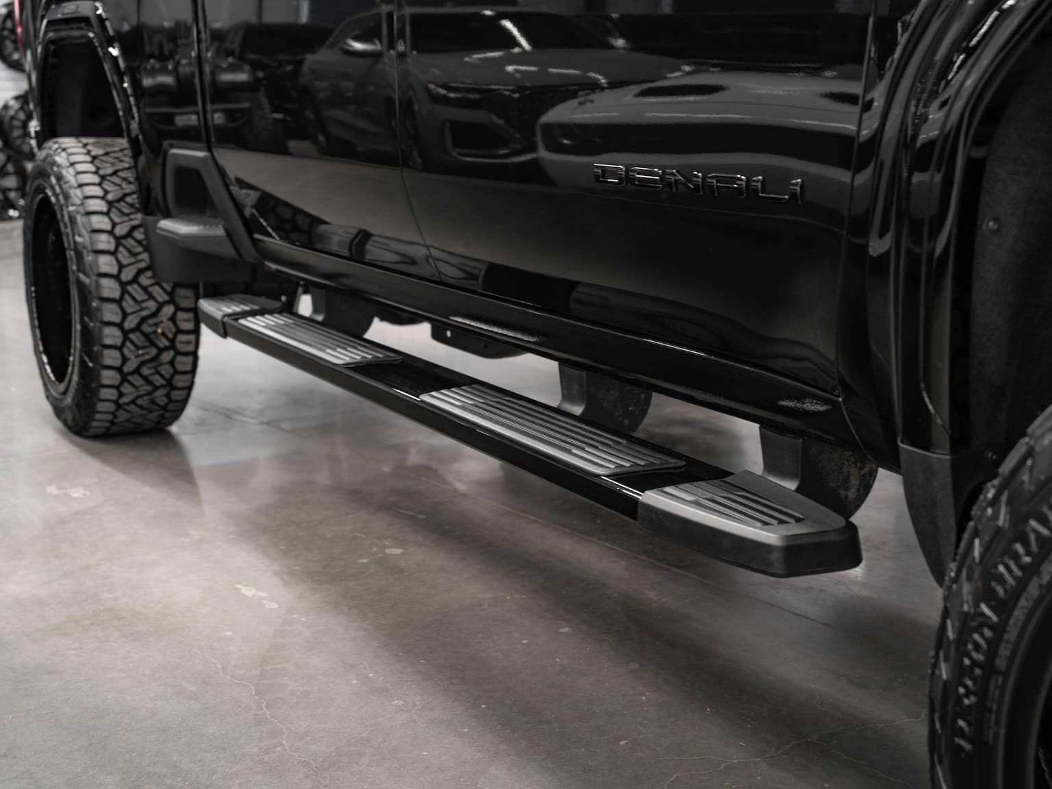 Black GMC Denali pickup truck featuring rugged tires and chrome side steps, parked on a polished concrete floor in a showroom atmosphere.