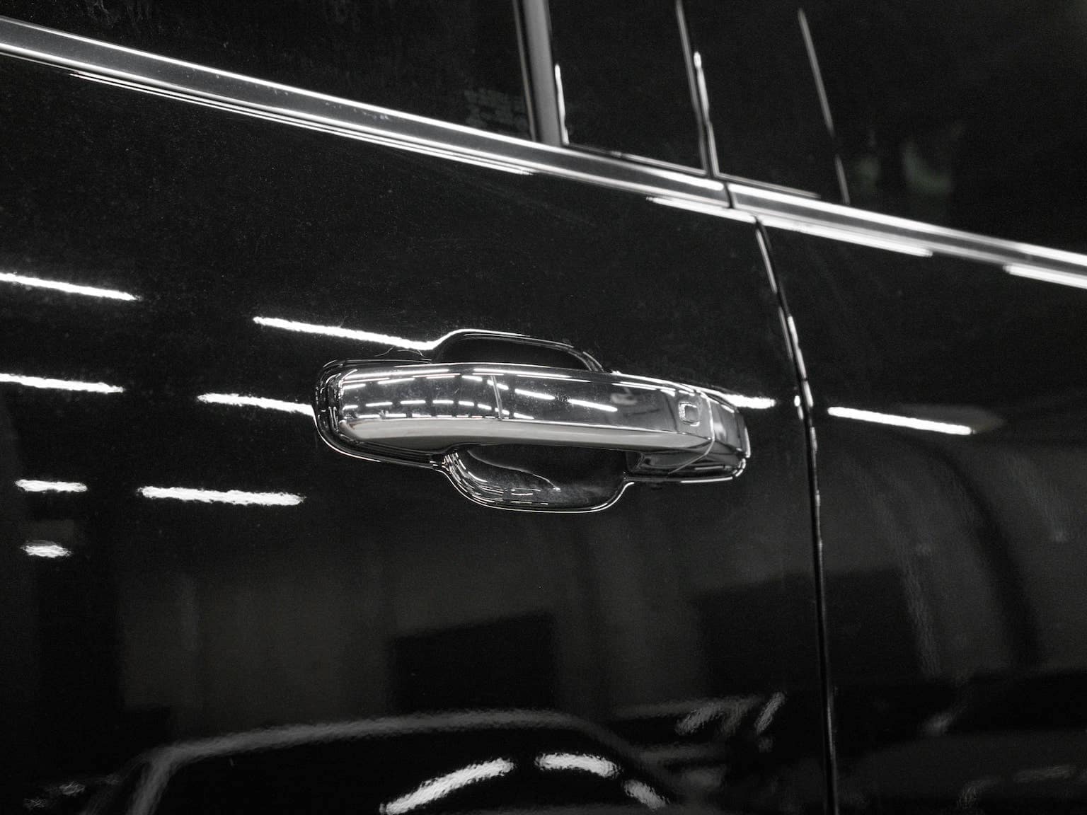 Close-up of a sleek chrome car door handle on a black vehicle, highlighting modern automotive design and reflective surfaces in a well-lit environment.