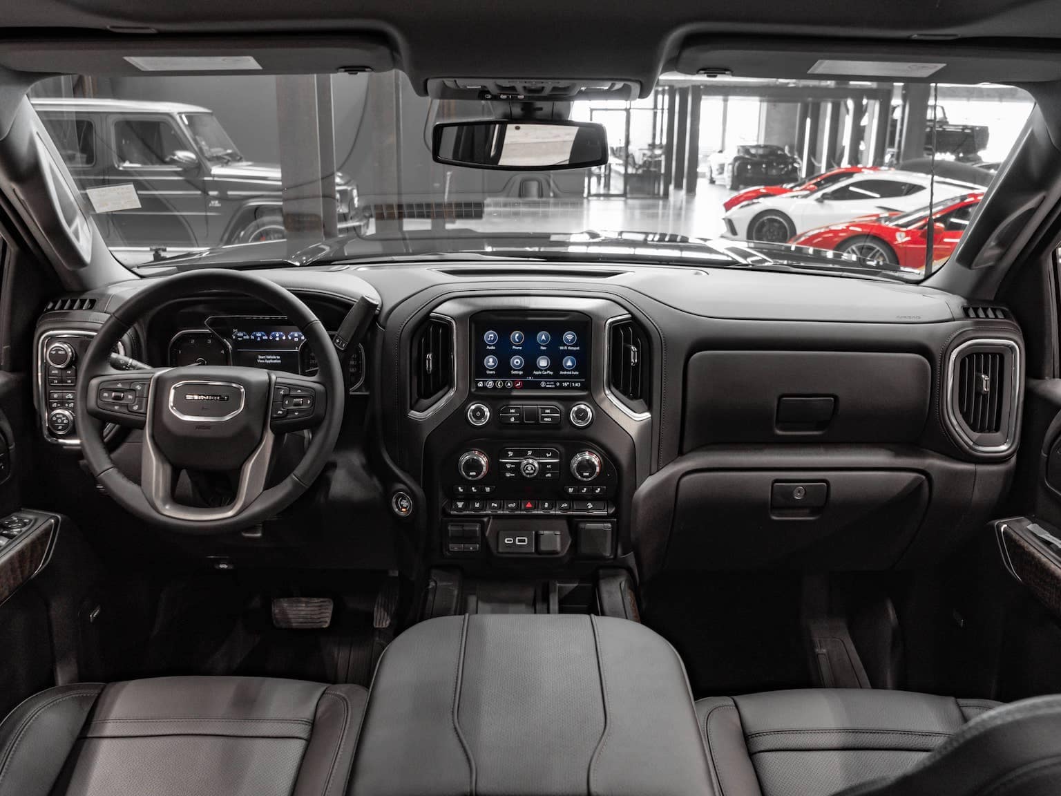 Interior view of a modern GMC truck featuring a sleek dashboard, advanced touchscreen display, and leather seating, set in a luxury car showroom with high-end vehicles in the background.