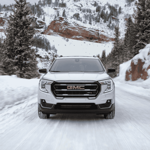 White GMC SUV driving on a snowy road surrounded by pine trees and rocky terrain, showcasing its performance in winter conditions.