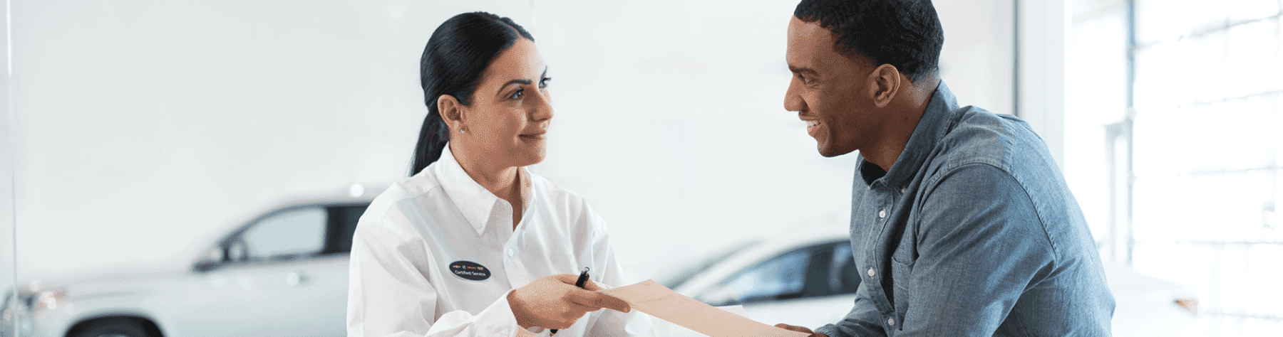 A customer service representative is handing paperwork to a male customer in a bright, modern dealership setting. The interaction represents a positive and engaging car buying experience.