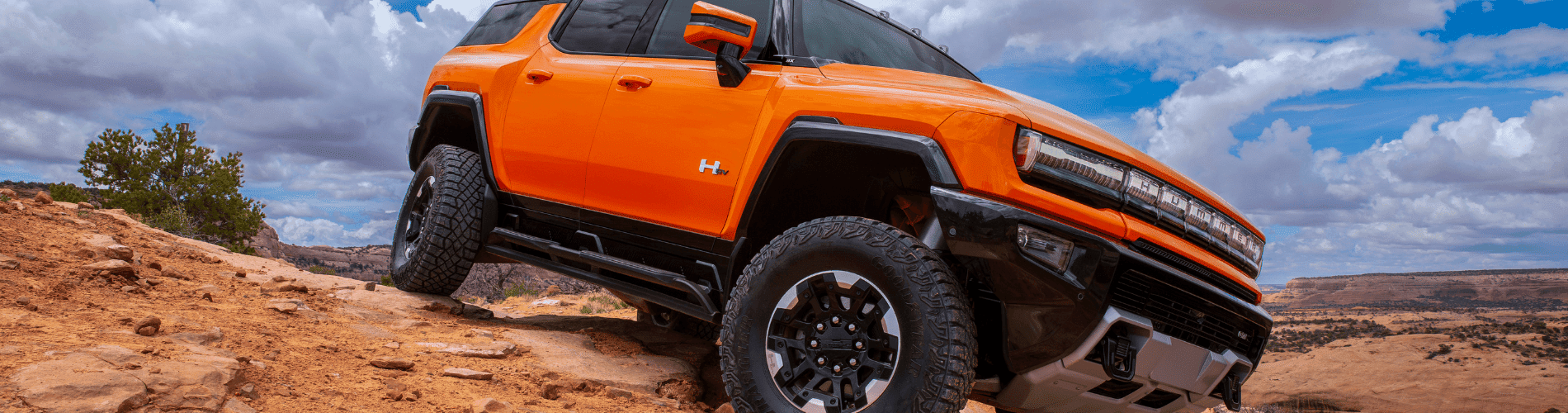 Bright orange electric Hummer SUV parked on rocky terrain under a scenic cloudy sky, showcasing its rugged design and off-road capabilities.