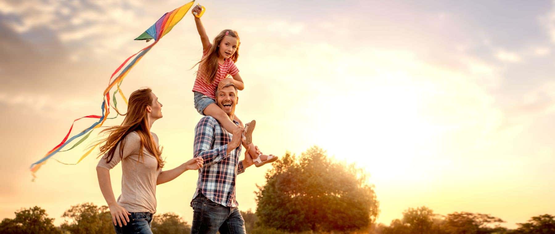 A happy family flying a kite during sunset, with a father carrying his daughter on his shoulders while the mother joyfully holds the kite string, surrounded by greenery.