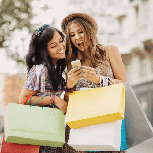 Two joyful women shopping outdoors, holding colorful shopping bags while looking at a smartphone.