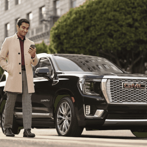 A stylish man in a beige coat checks his smartphone next to a black GMC Denali SUV parked on a city street, showcasing luxury and modern design.