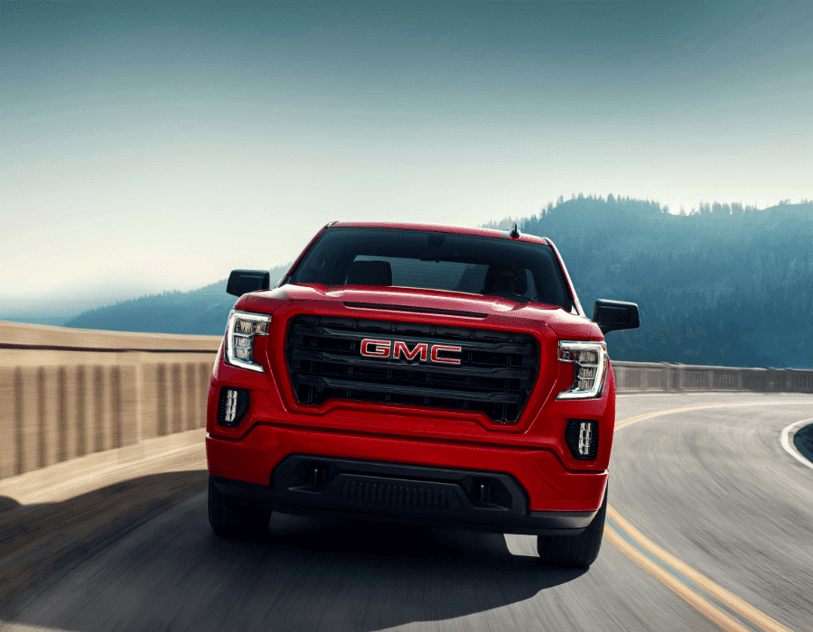 Red GMC truck driving on a scenic winding road with mountains in the background, showcasing its bold front grille and sleek design. Perfect for off-road adventures and daily commutes.