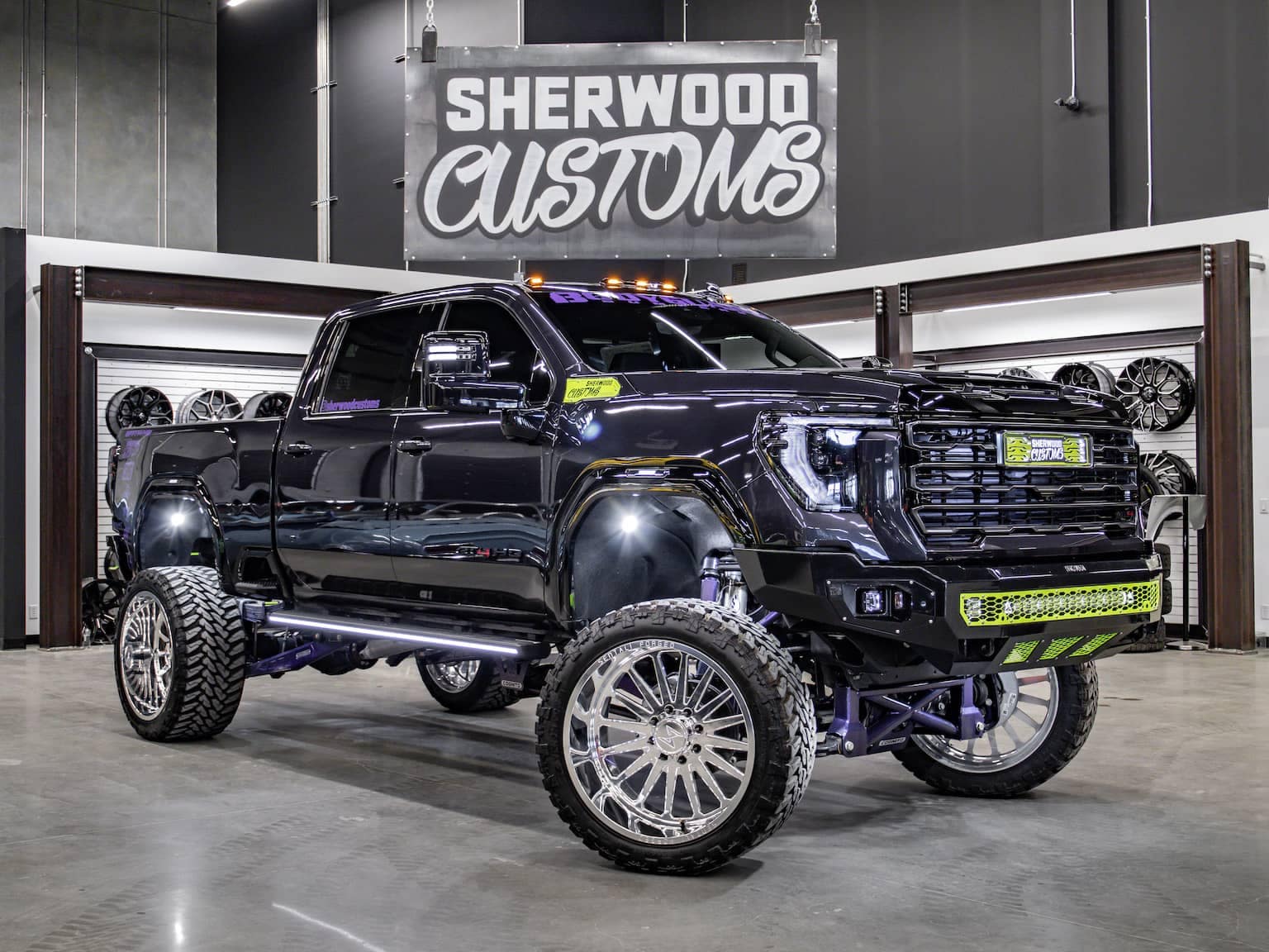 Custom lifted black truck with oversized wheels and eye-catching neon accents, showcased in a modern auto shop setting featuring custom rims and a prominent "Sherwood Customs" sign in the background.