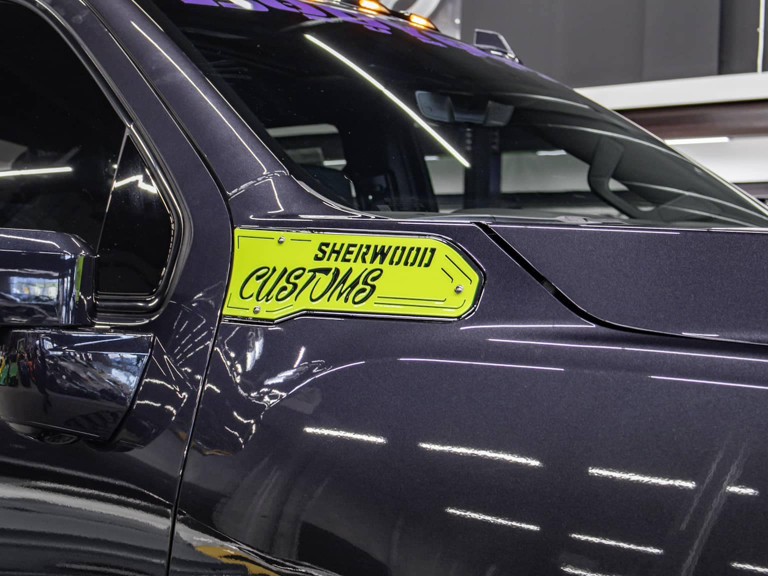 Close-up of a custom vehicle showcasing a vibrant yellow "Sherwood Customs" decal on the side. The image highlights the glossy dark exterior and detailed design elements of the vehicle, emphasizing its unique modifications and custom features.