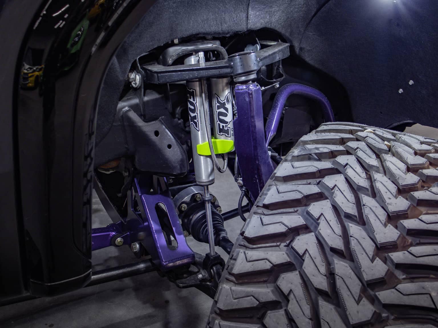 Close-up of a truck's suspension system, featuring Fox Racing shocks, purple control arms, and a rugged tire tread, showcasing high-performance off-road capabilities.