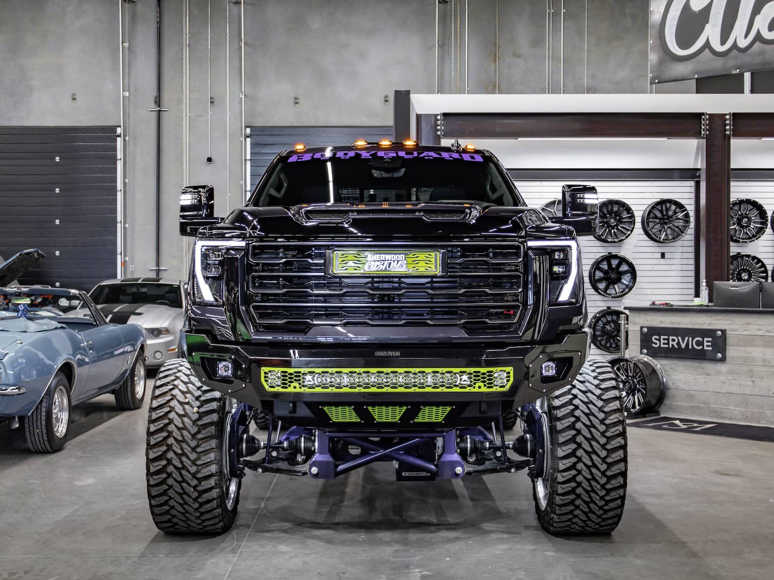 Custom lifted black truck with oversized tires and an aggressive front grille on display in a automotive shop. The background features classic cars and wheel displays, emphasizing the truck’s rugged design and performance enhancement.