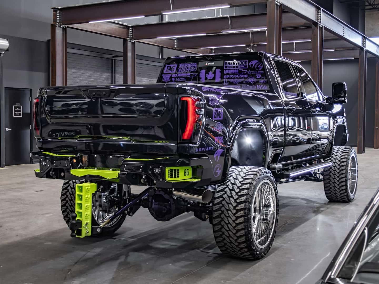 Rear view of a customized black pickup truck featuring a raised suspension, oversized wheels, and neon green accents, showcased in a modern indoor showroom setting.