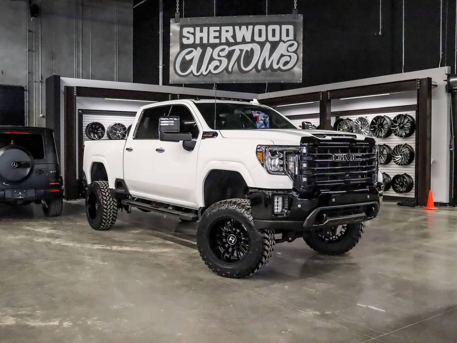 Custom GMC truck displayed at Sherwood Customs, featuring a lifted suspension, off-road tires, and a sleek white exterior. The backdrop includes a wall of custom wheels, emphasizing the customization services offered.